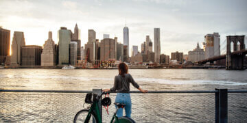 Individual standing by a railing with a bicycle, gazing at the New York City skyline across the water.